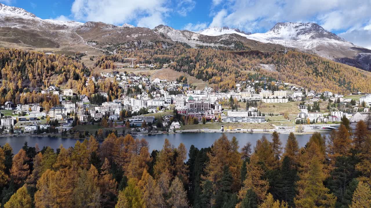 An aerial showing the luxury resort St. Moritz Dorf nestled between Lake St. Moritz and golden larch forests. The high-altitude town is backed by snow-dusted peaks under a clear, autumnal sky