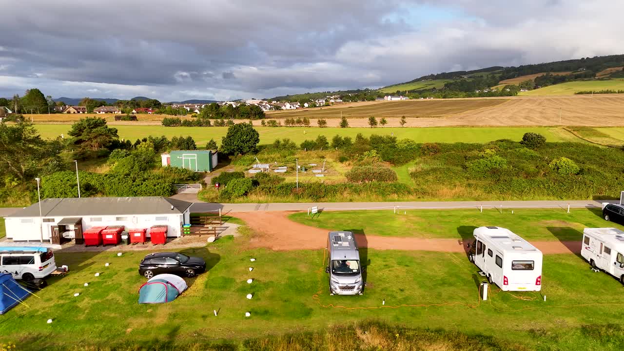 Drone footage moves forward above a rural caravan and camping site with motorhomes, tents, and vehicles under partly cloudy daylight in the Scottish Highlands