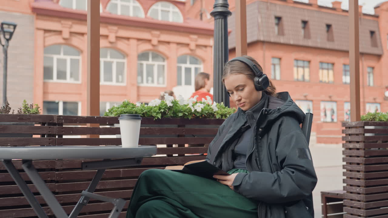 Женщина в наушниках читает что-то на планшете на улице, на скамейке в городе; на заднем плане — здания из красного кирпича, на перилах — горшечные растения, мягкий дневной свет, повседневная куртка, большие наушники, прокрутка