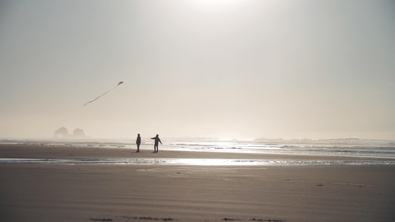 People flying a kite together on an empty beach at the ocean while sunset with rocks in the background - relaxing activity - 4k slow motion