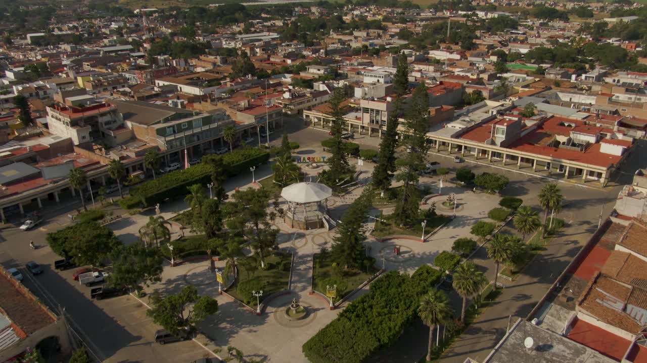 Upward aerial from central kiosko revwals symmetrical park layout, palm-lined streets, and surrounding town buildings in Tuxpan, Jalisco