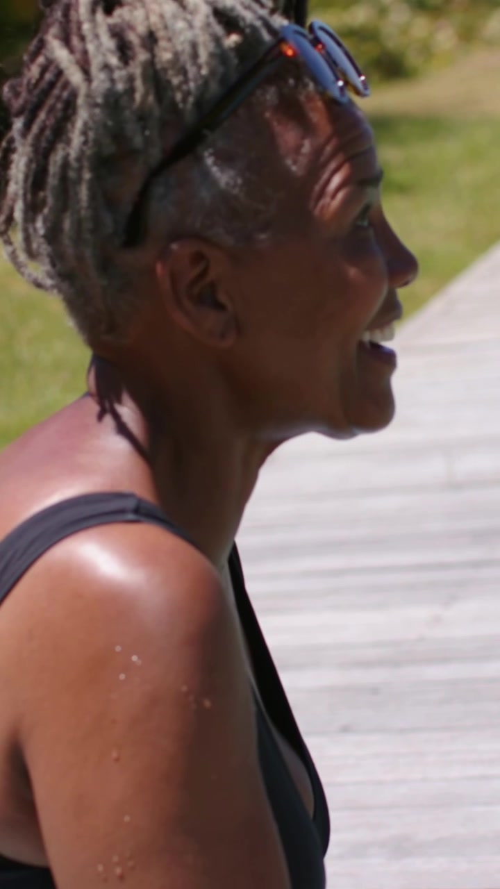 Vertical video: Smiling woman in black swimsuit lifting glass after off-camera call on wooden dock