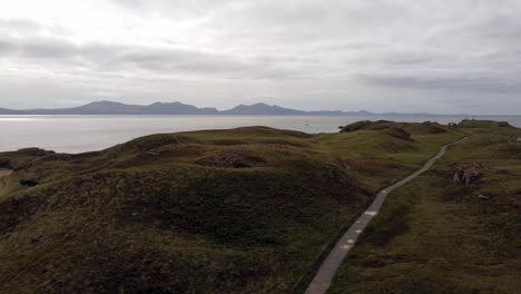 Aerial-view-Ynys-Llanddwyn-island-Anglesey-coastal-walking-trail-with-Snowdonia-mountains-across-the-Irish-sea-orbiting-left