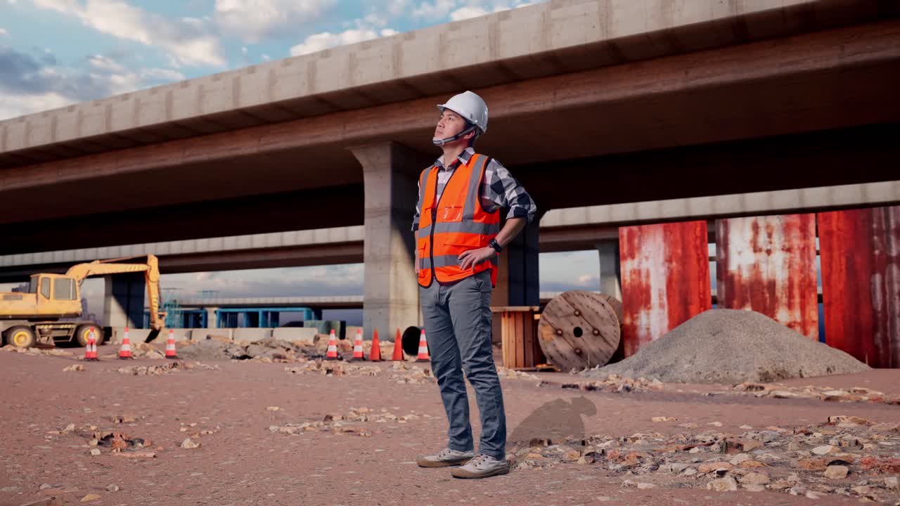Full Body Side View Of Asian Male Engineer Wearing Safety Helmet Looking Around While Standing With Arms Akimbo At The Site of Highway Construction
