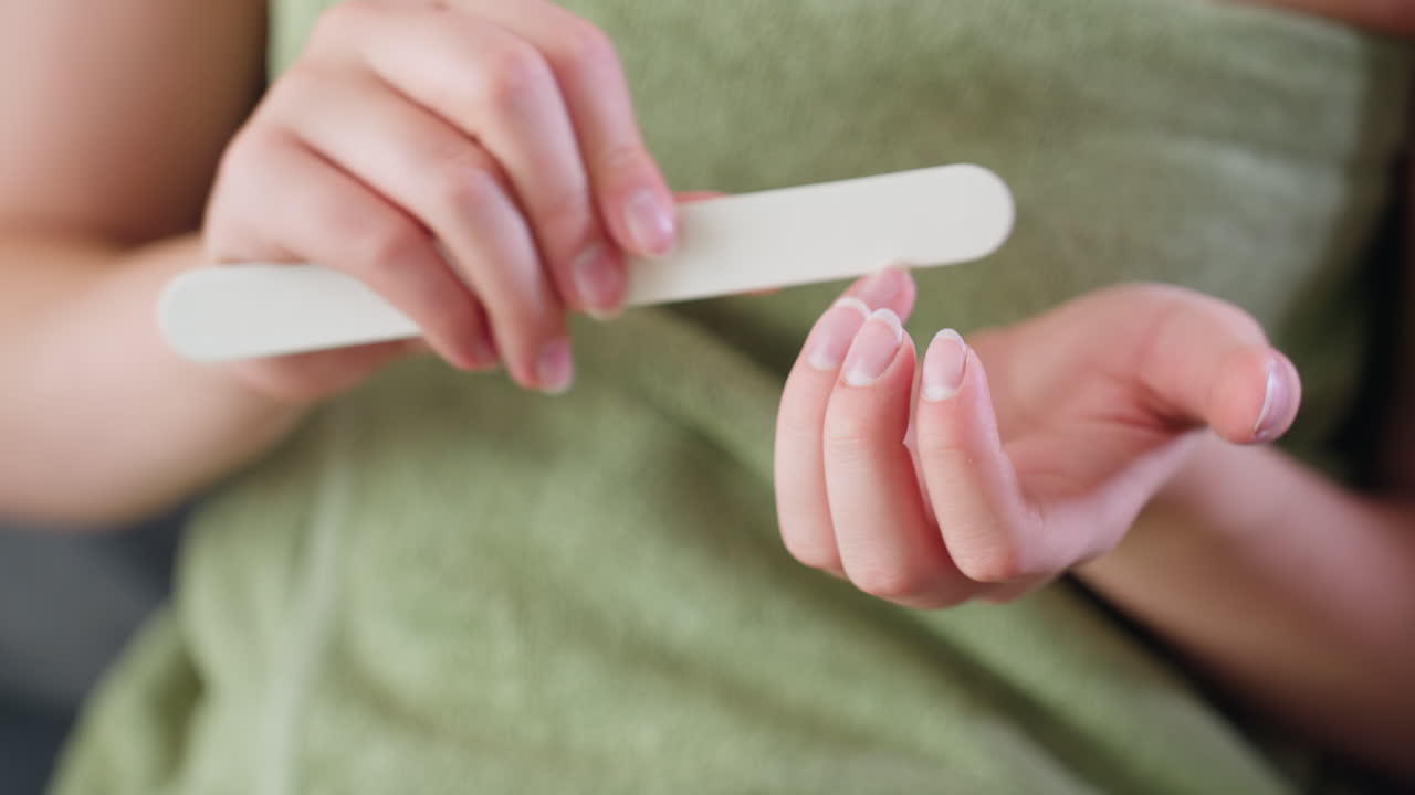 Close up view of woman s hands gently filing nails while wrapped in green towel, fine nail dust visible in motion, indoor setting with natural lighting, expressing calm grooming and personal care