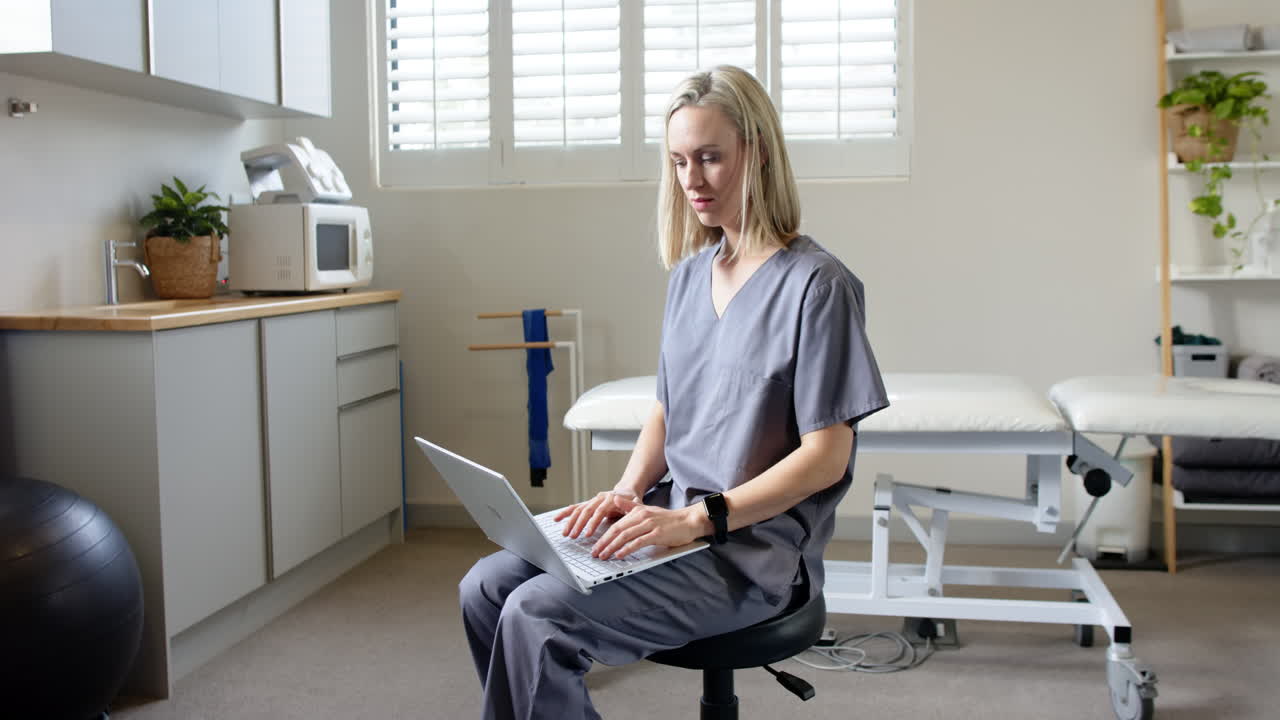In prosthetic lab, woman in scrubs using laptop while sitting on stool