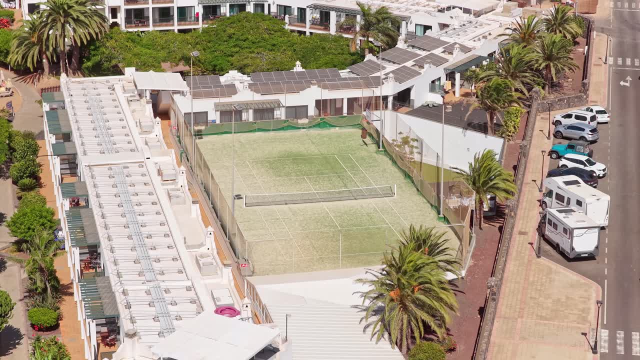 Corralejo’s tennis court zone with fenced court, palm-lined buildings, solar rooftops, and parked vehicles showing sport and residential planning in northern Fuerteventura