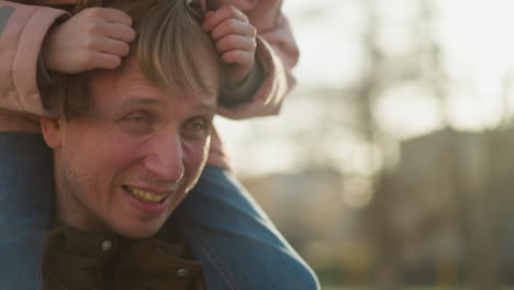 cheerful father smiles as his little girl, perched on his shoulders, playfully arranges his hair in the warm light of sunset