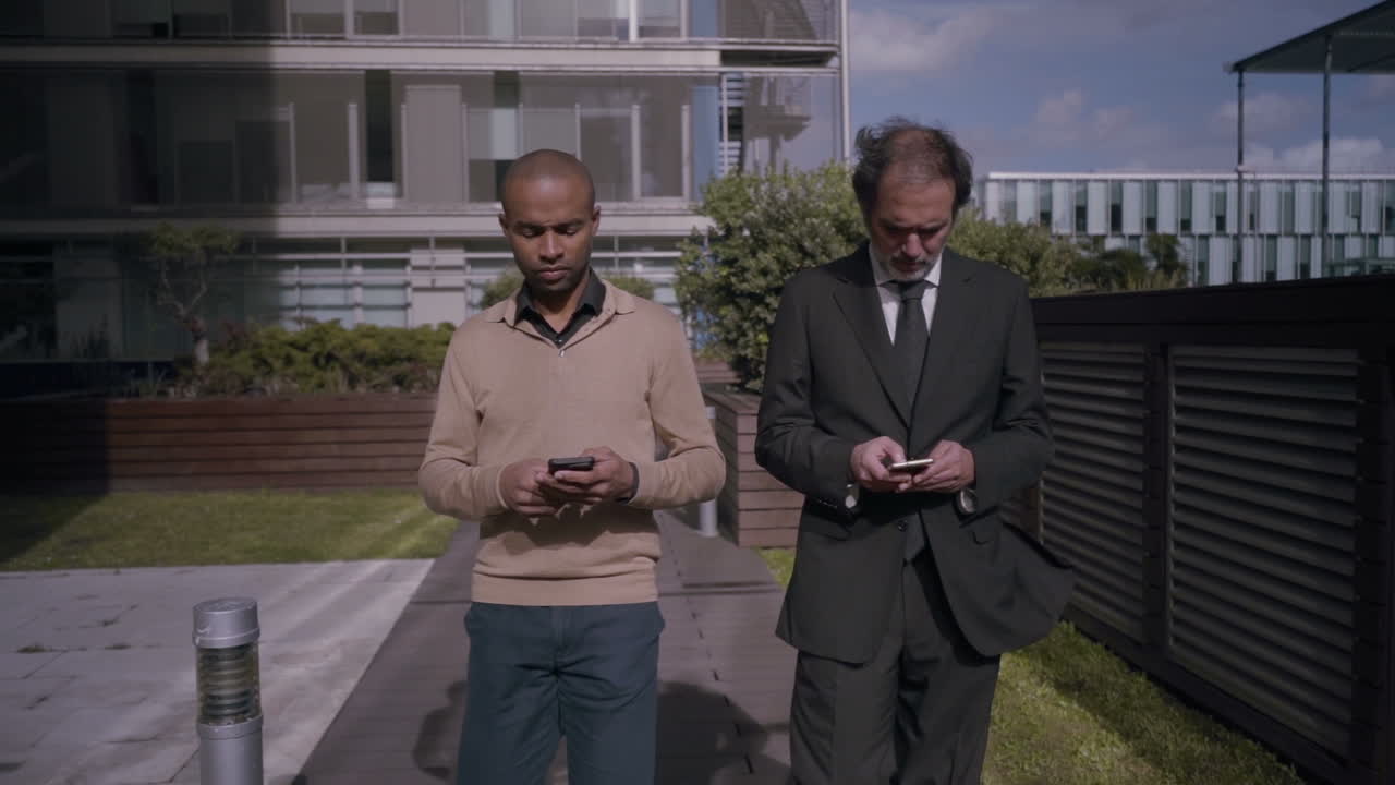 Empresarios concentrados caminando y usando smartphones cerca de edificios de oficinas