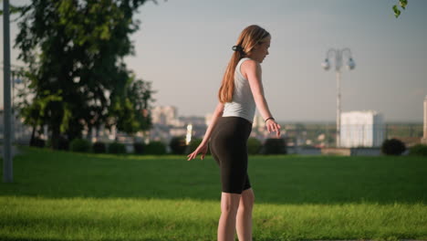 side view of young white woman skating outdoors, swinging her hands wide open for balance on sunny day with blurred cityscape in background, surrounded by lush greenery, lamp post, and trees