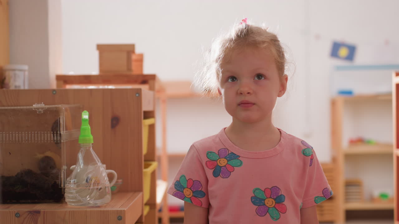 Little girl in kindergarten with braided hair and pink shirt stands quietly trying to maintain focus under sunlight, wooden shelves and classroom background