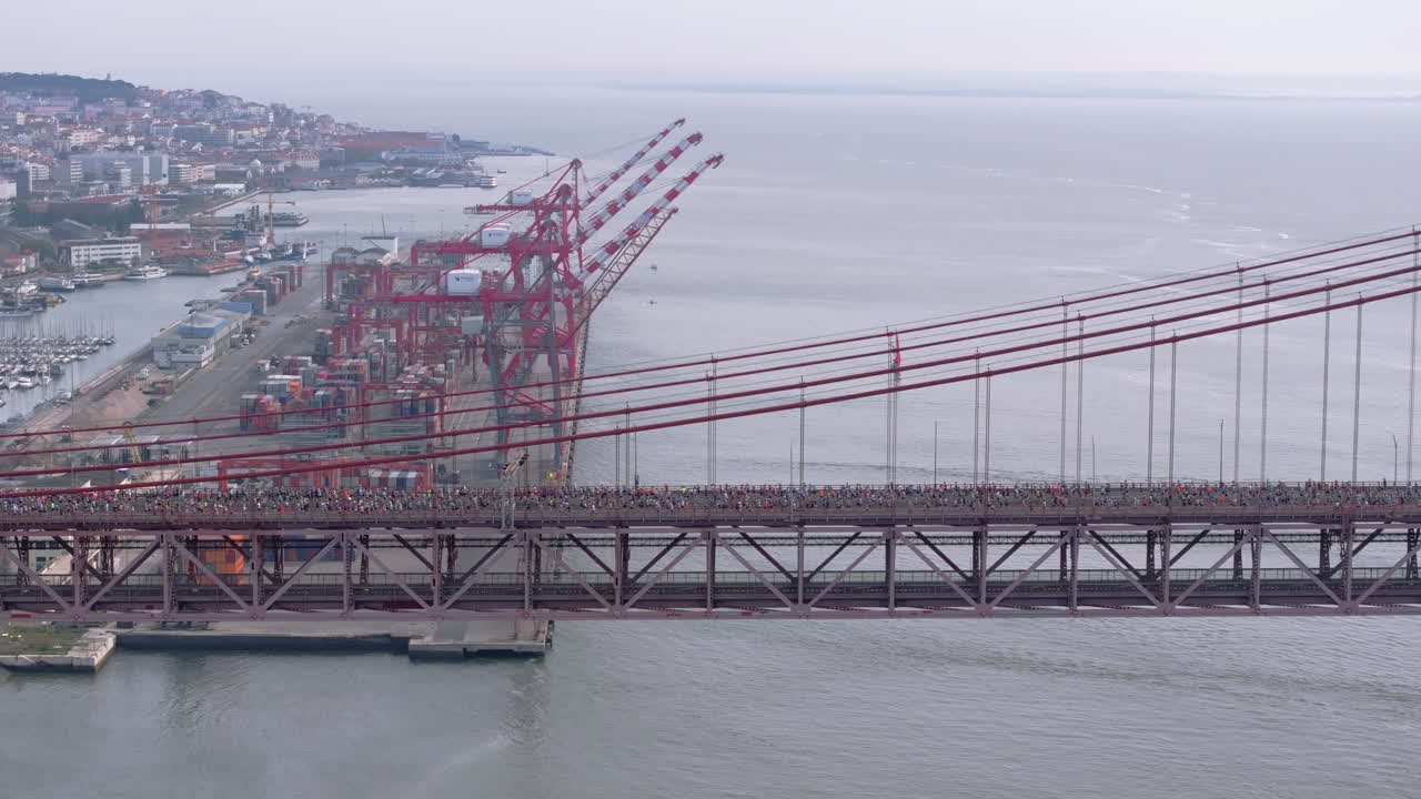 Side-on aerial drone shot of half marathon and 10K long-distance running event in Lisbon, Portugal, Europe. Runners crossing the iconic famous red 25th April bridge by Alcantara docks
