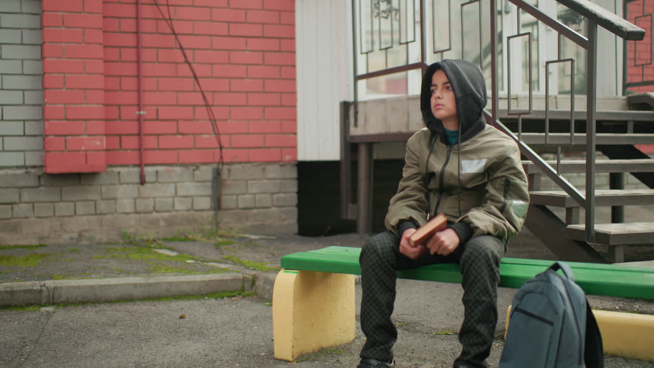 Young kid in hooded jacket seated on outdoor bench holding and tapping book in left hand, backpack placed beside, thoughtful expression against brick wall and staircase background
