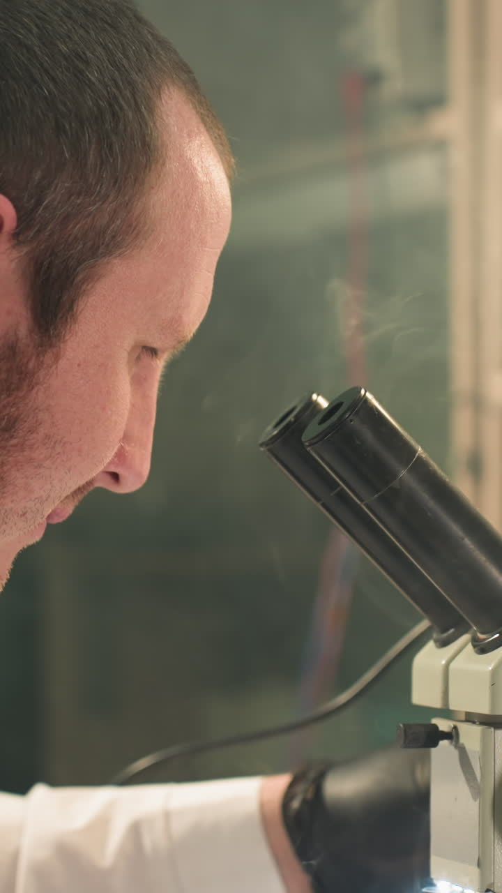 una vista de cerca de un técnico en una bata de laboratorio examinando atentamente una placa de circuitos electrónicos bajo un microscopio, el espacio de trabajo está iluminado por una lámpara de escritorio, con humo visto