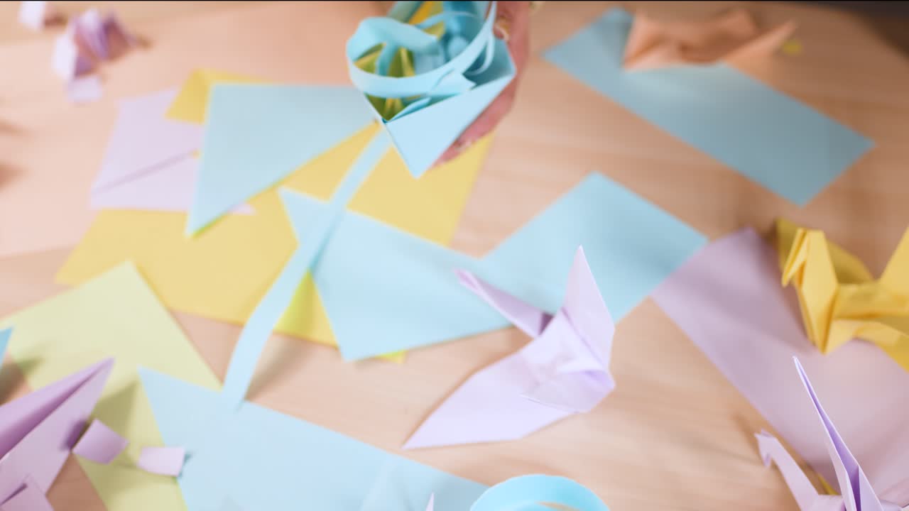 Overhead view of hands meticulously arranging pastel origami paper crafts on a wooden work table