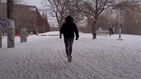 man wearing parka and boots running on snow covered ground toward park between buildings in montreal
