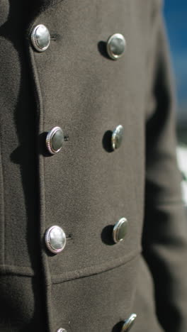 primer plano de un elegante abrigo negro con botones metálicos brillantes en un entorno urbano al aire libre con fondo borroso, incluidos coches estacionados y vegetación