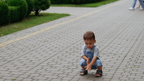 cute baby boy in jeans romper squats on the paved road. interested kid touches the tiles on the ground.