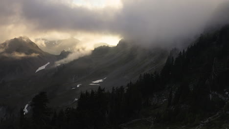 Golden-hour-light-diffused-between-thick-clouds-above-silhouette-of-evergreen-PNW-forest-below-rocky-slopes