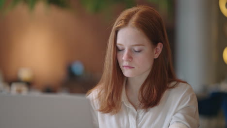 thoughtful brooding remote working red haired woman sitting infront of a laptop or notebook in casual outfit on her work desk in her modern airy bright living room home office with many windows