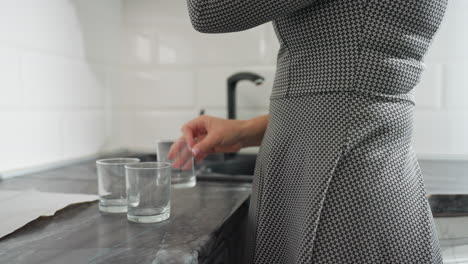 partial view of lady in plaid gown standing in kitchen placing three transparent glass cups carefully on countertop near sink in bright clean space with natural sunlight streaming through curtains