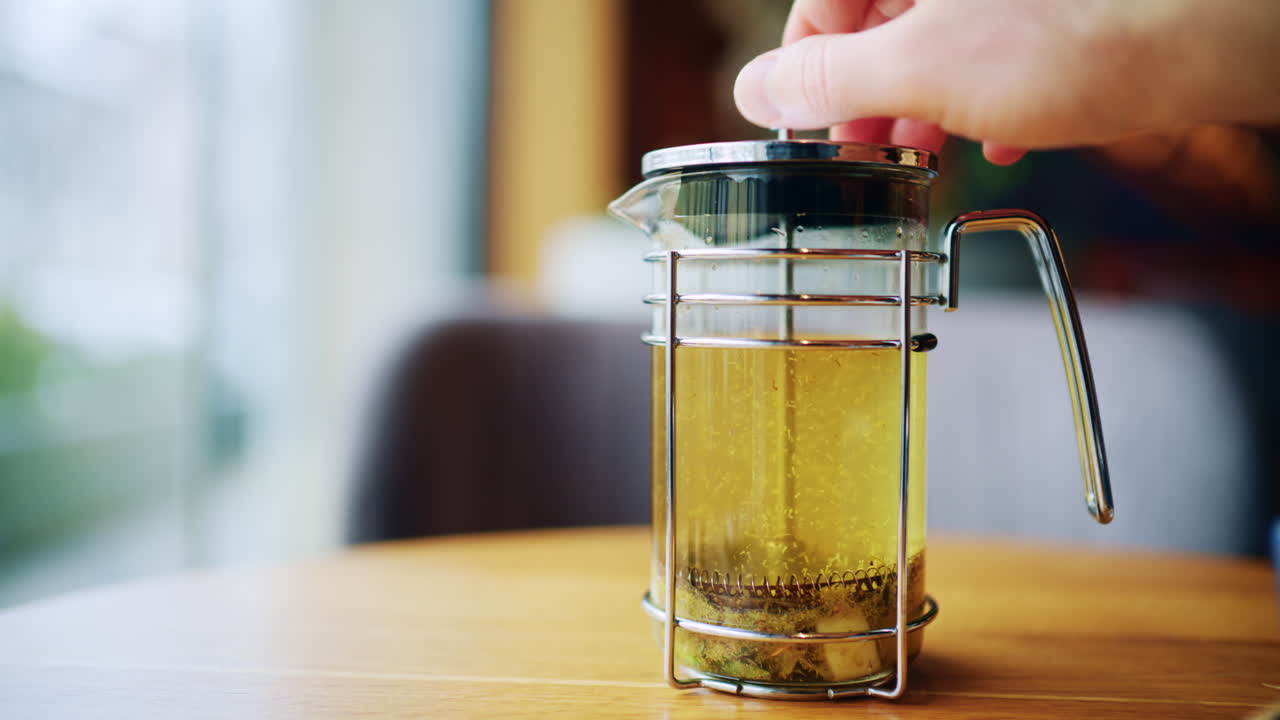 Donna che preme una French press per preparare una tisana in una teiera di vetro su un tavolo di legno in un ambiente accogliente.