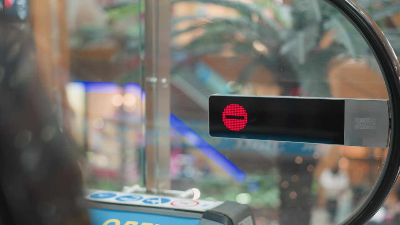 red stop indicator display mounted on glass panel of moving walkway while shopper steps off onto mall corridor with blurred pedestrian and vibrant store front lighting in modern mall background