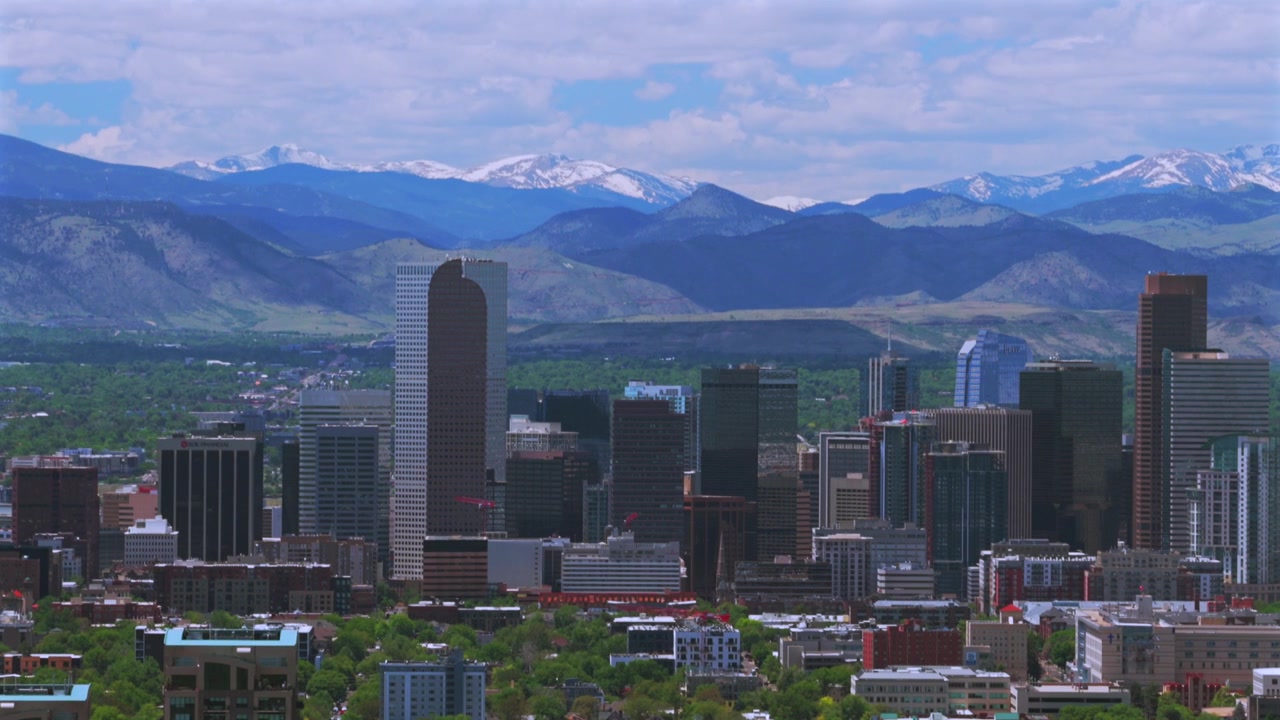 Ripresa aerea con drone di Denver, la "Mile High City", Colorado, con le Montagne Rocciose della Front Range innevate, lo skyline di grattacieli e gli edifici del centro. Mattina di primavera con cielo azzurro e nuvole, movimento circolare a sinistra.