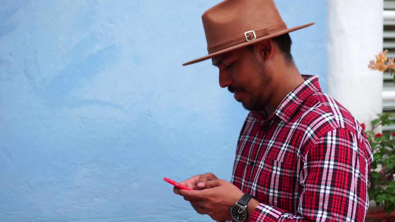 hombre colombiano positivo usando teléfono móvil de pie contra la pared azul