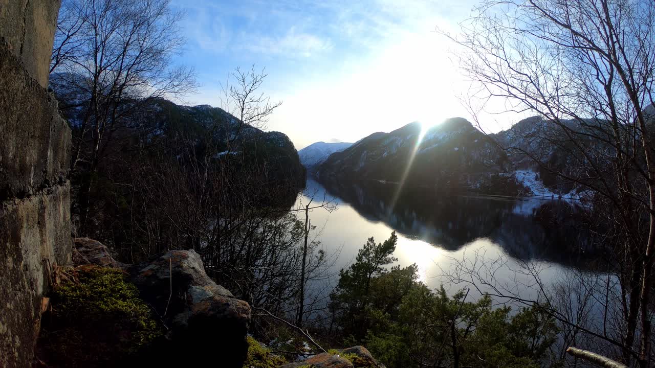 ノルウェーのヴェフィヨルドに沈む広角の夕日 – オステロイ島の山の後ろに太陽がゆっくりと沈む間に通過する雲 – 前景に茂みや木々が海に反射