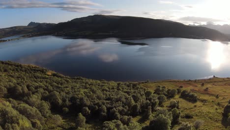 Entlang-Eines-Berges-Hinunter-Zum-See-Mit-Blick-Auf-Die-Graslandschaft-Und-Die-Vielen-Bäume-Am-Hang-Fliegen,-Dann-In-Zeitlupe-über-Den-See-Mit-Seiner-Glatten-Oberfläche-Fliegen