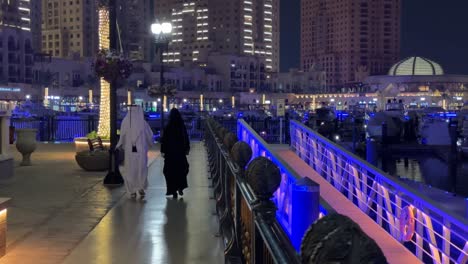 Arab-couple-man-with-traditional-white-long-tidy-clean-dress-and-woman-with-black-long-dress-Islamic-hijab-are-walking-at-night-in-a-luxury-scenic-wonderful-city-landscape-in-Pearl-Doha-Qatar-island