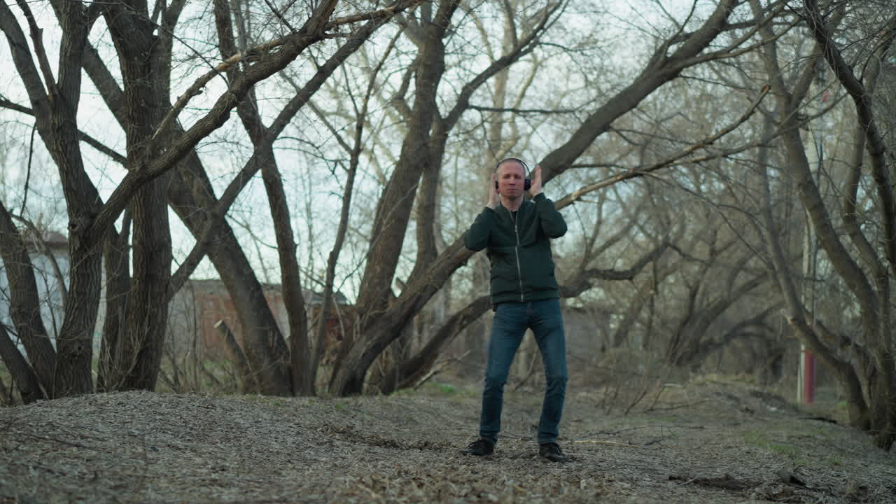 녹색 재을 입은 남자가 조용한 자연 환경에서 그의 움직임을 포착하여 조용한 숲에서 헤드폰으로 춤을 춘다