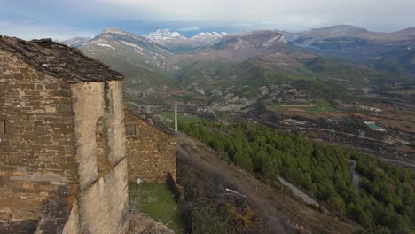 Disparo-De-Drone-Desde-Una-Iglesia-Abandonada-En-Muro-De-Bellos,-España,-Con-Un-Hermoso-Paisaje-Verde-Y-Una-Montaña-Nevada-Como-Fondo