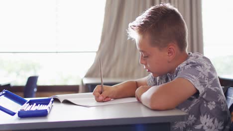 Portrait-of-happy-caucasian-schoolboy-sitting-at-classroom,-making-notes,-looking-at-camera