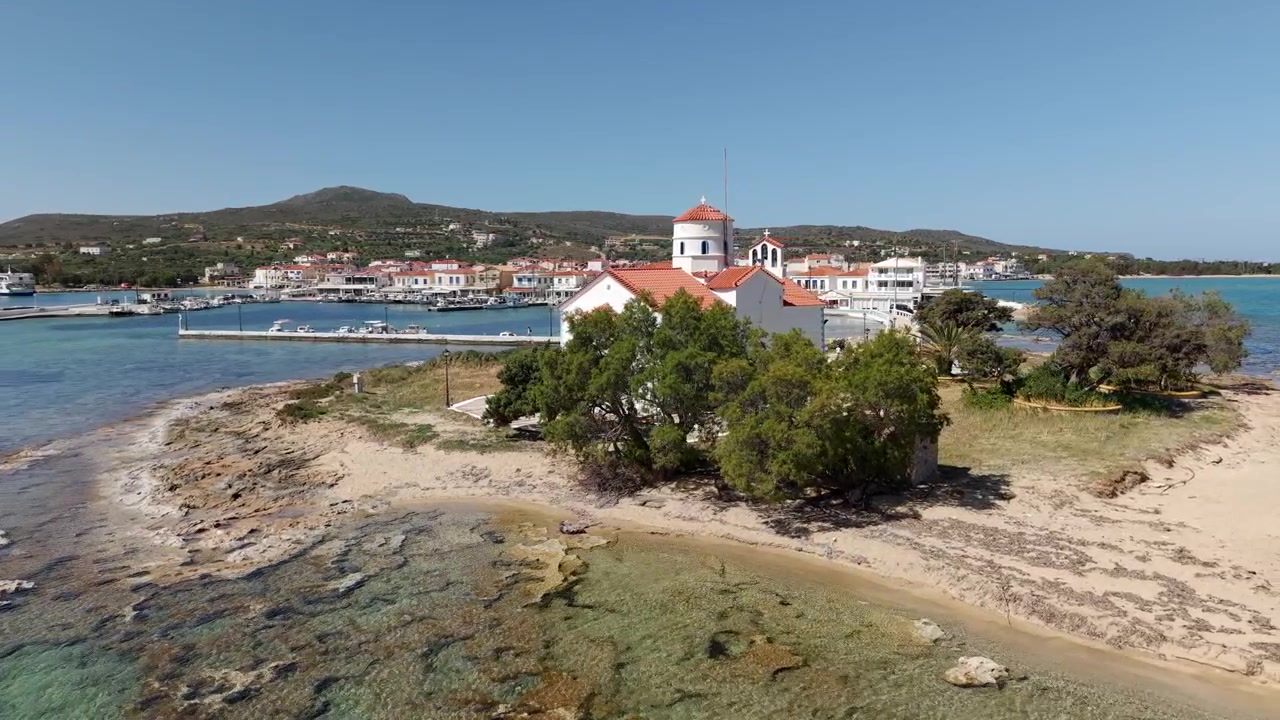 Drone circula a antiga igreja caiada de Agios Spyridon na costa de uma pequena ilha rochosa em um dia ensolarado, casas à beira-mar da vila de Elafonisos ao longo do porto principal através do mar Jônico