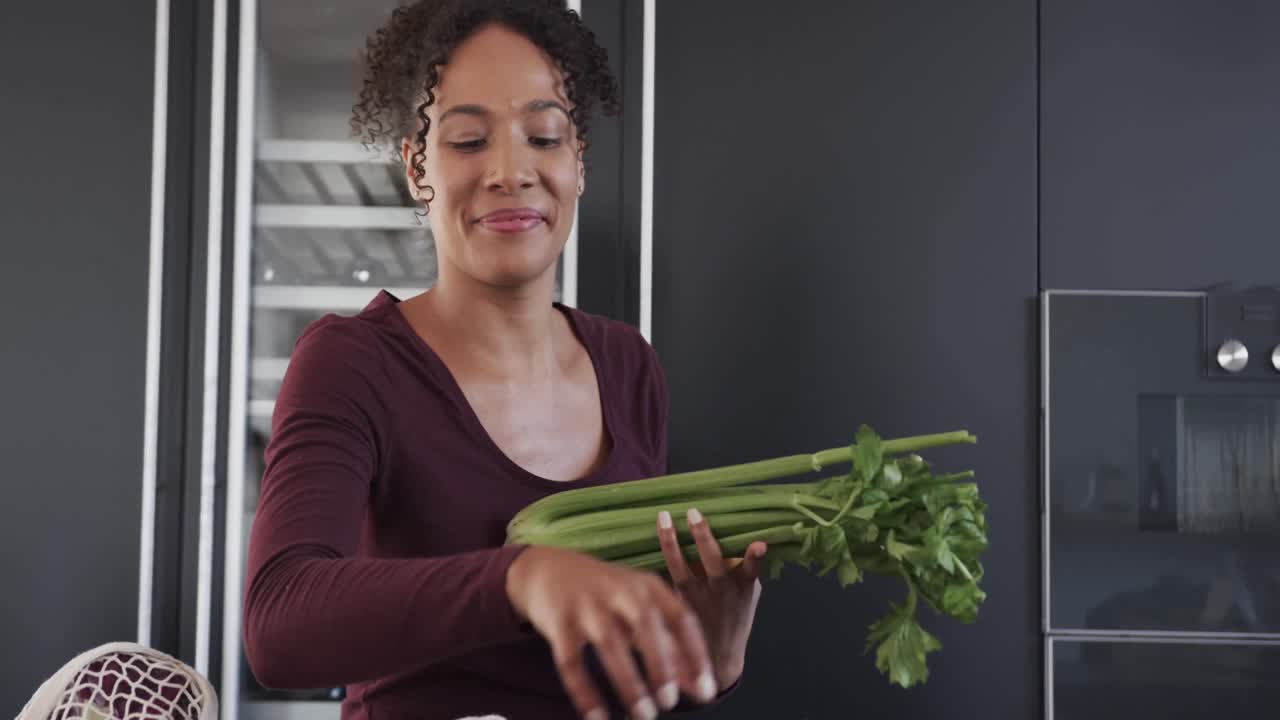 feliz mujer biracial poniendo bolsas de compras de comestibles y sonriendo en la cocina, en cámara lenta