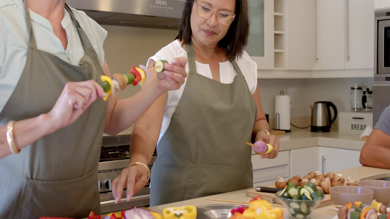 Preparing vegetable skewers, senior diverse women cooking together in modern kitchen, at home