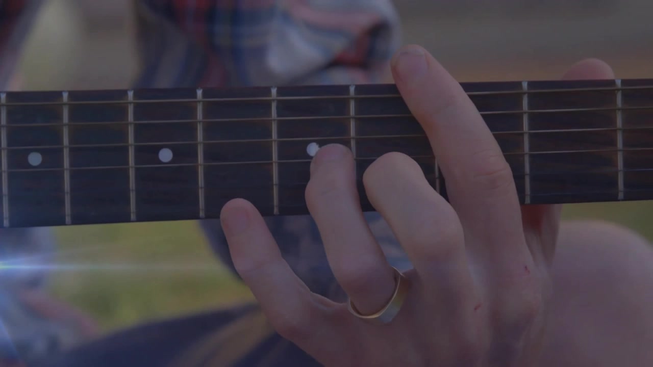 Vänster hand trycker ner ackord och glider sedan fingret för nästa ackordbyte i en solig musikhandledning utomhus.