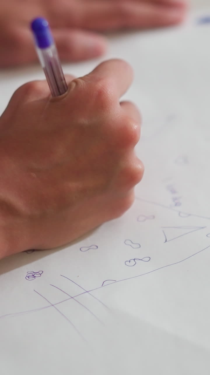 close up of kid wearing red rope around wrist sketching on white paper laid on floor using blue pen with simple drawings and playful shapes expressing early creativity focus