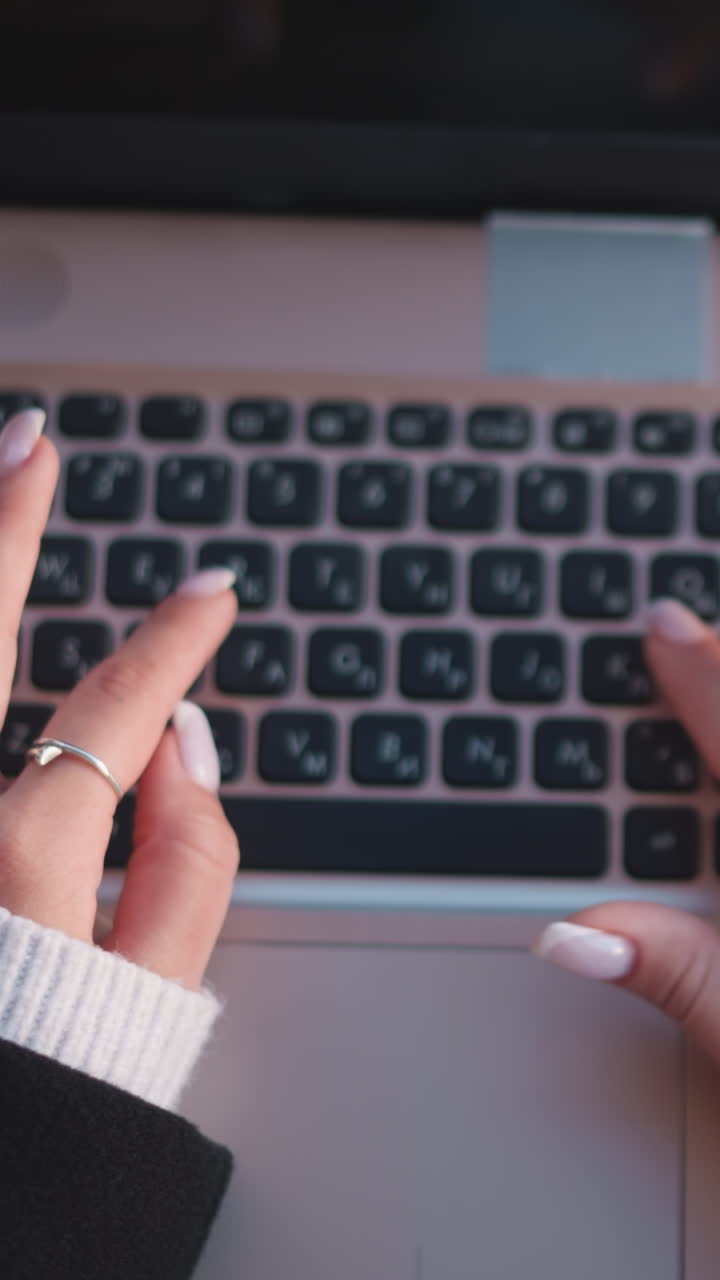 primer plano de manos con uñas pulidas escribiendo en un teclado de portátil, adornado con elegantes anillos, cómodas mangas de suéter y un teléfono inteligente en una mesa de madera
