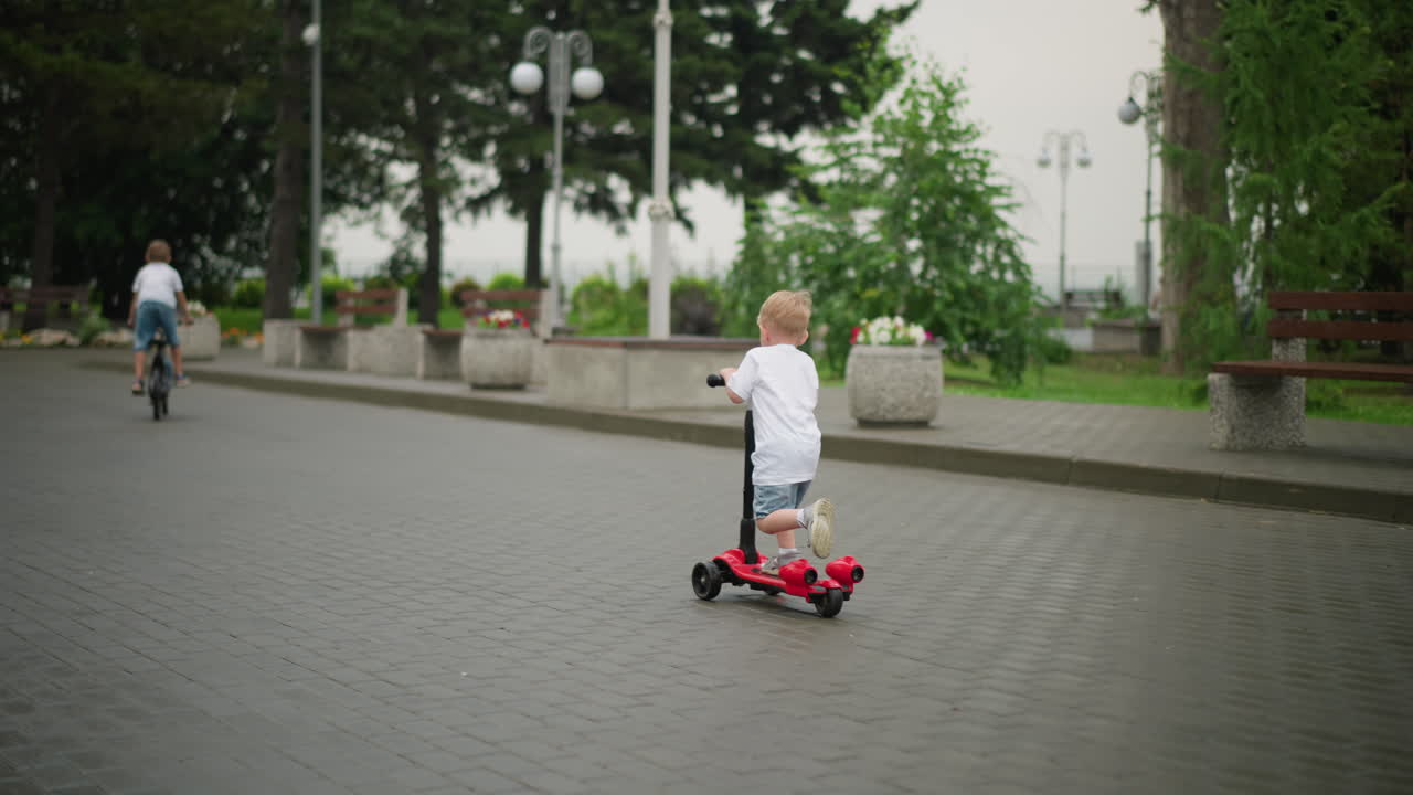 un niño pequeño monta un scooter rojo en un amplio parque, con una camisa blanca y pantalones cortos de vaqueros, con otro niño montando una bicicleta delante, con bancos vacíos, plantas en maceta y árboles alrededor