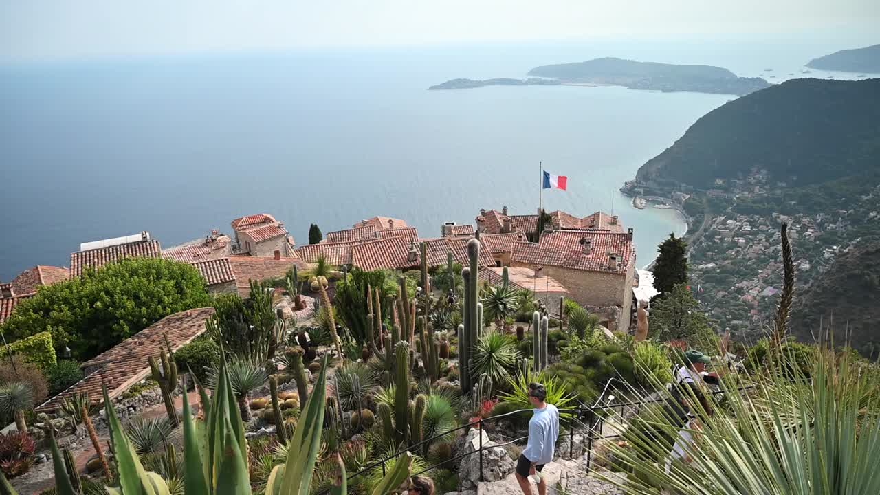 MENTON, FRANCE - SEPTEMBER, 2021: Park with tropical plants. People, roofs of traditional residential buildings, sea on the background