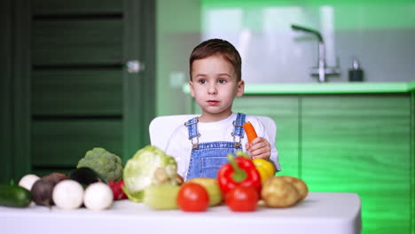 a range of diverse vegetables on the table in front of the little boy. cute toddler is eating a carrot in the kitchen.