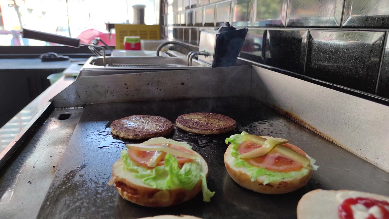 業務用の鉄板でジュージューと音を立てて焼けるビーフバーガーパティとトッピングされたバンズ。