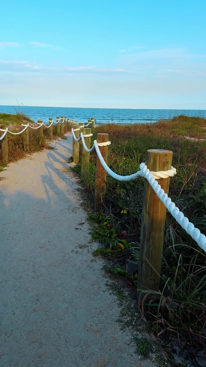 Sentiero sabbioso con ringhiera di corda che porta alla spiaggia dell'oceano. Passerella costiera tra dune di sabbia con pali di legno e recinzione di corda bianca che si dirige verso il mare blu.