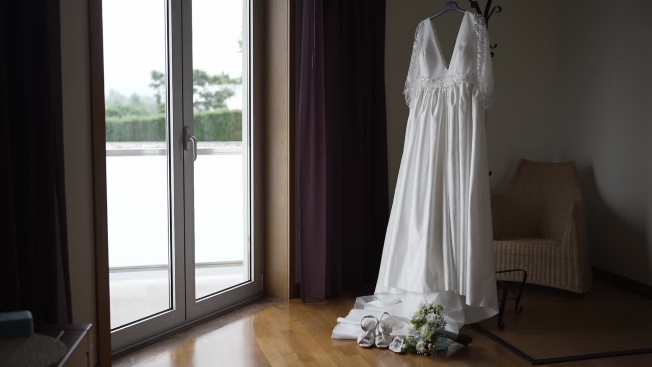 White wedding dress hanging near a window with bridal accessories on the floor