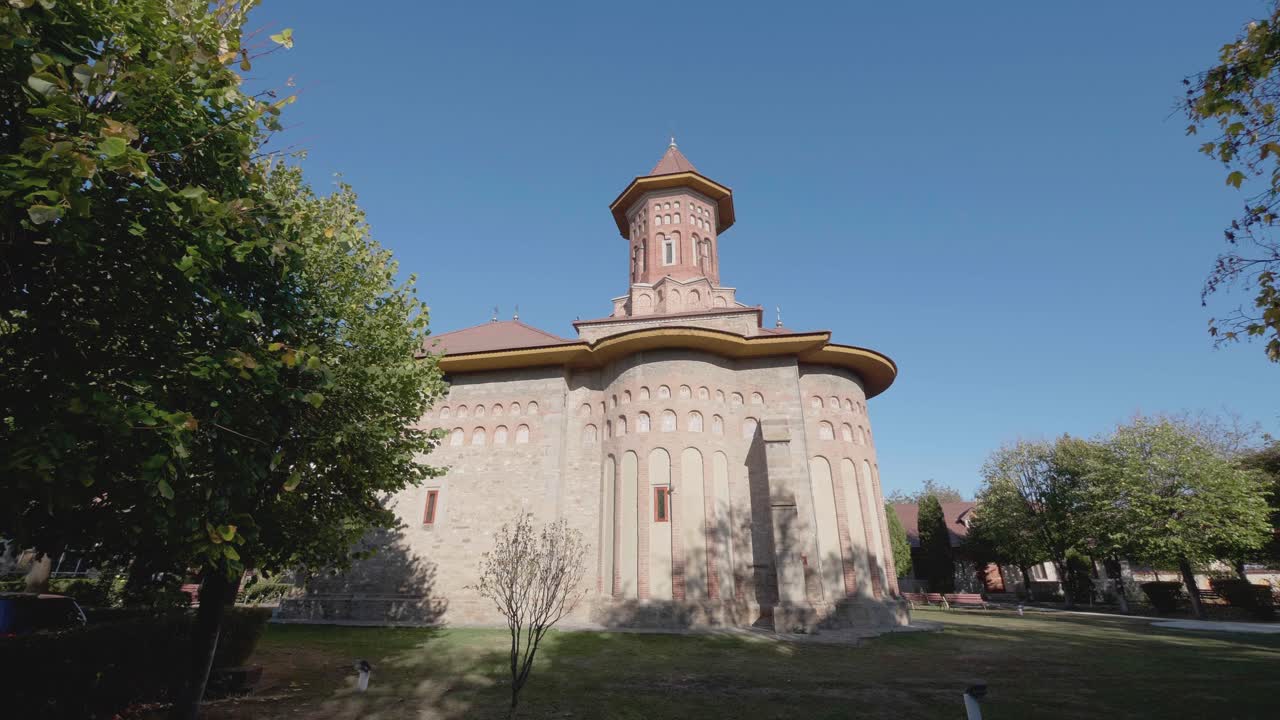 A low-angle tilting shot reveals the historic Precista Church in Bacau, Romania. The camera moves up the medieval stone and brick walls to the tower against a clear blue sky