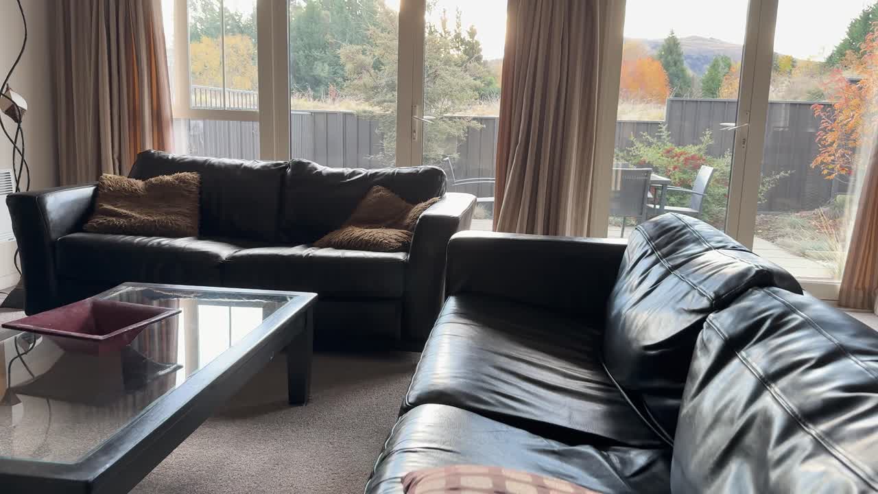 Camera smoothly moves into a sunlit living room with large windows, leather sofas, glass coffee table, and neutral decor, highlighting a cozy, inviting home interior