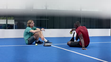 diverse men sitting on padel indoor court, discussing game strategy with rackets in hand, copy space